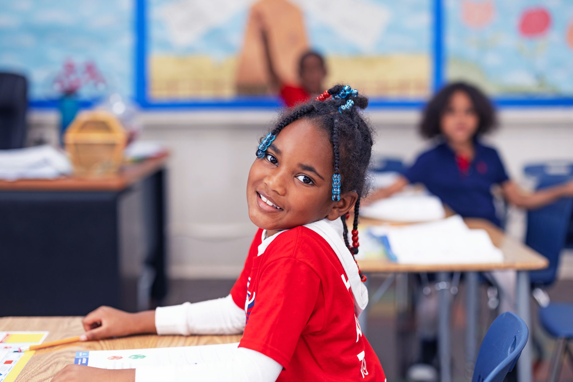 Smiling young student with beaded braids at a classroom desk.