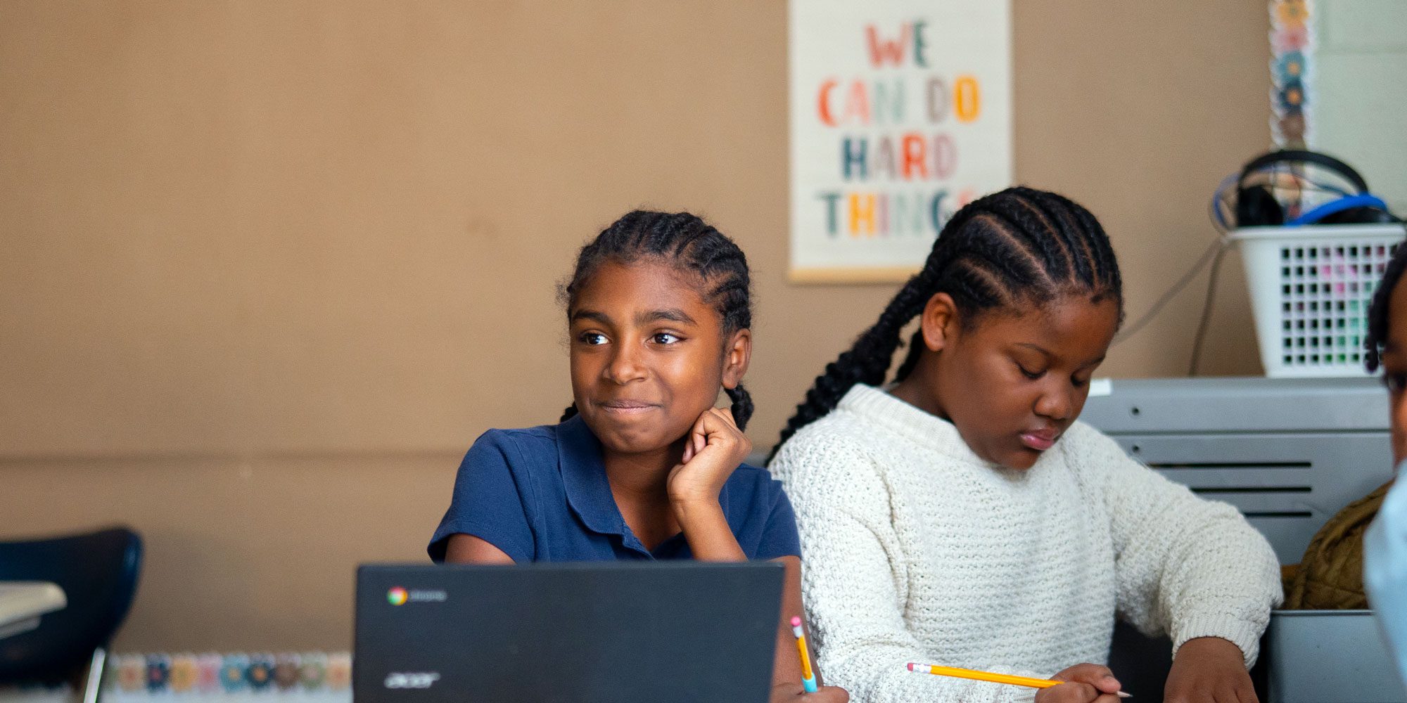 Student at a laptop beside a classmate writing under a "We Can Do Hard Things" poster.
