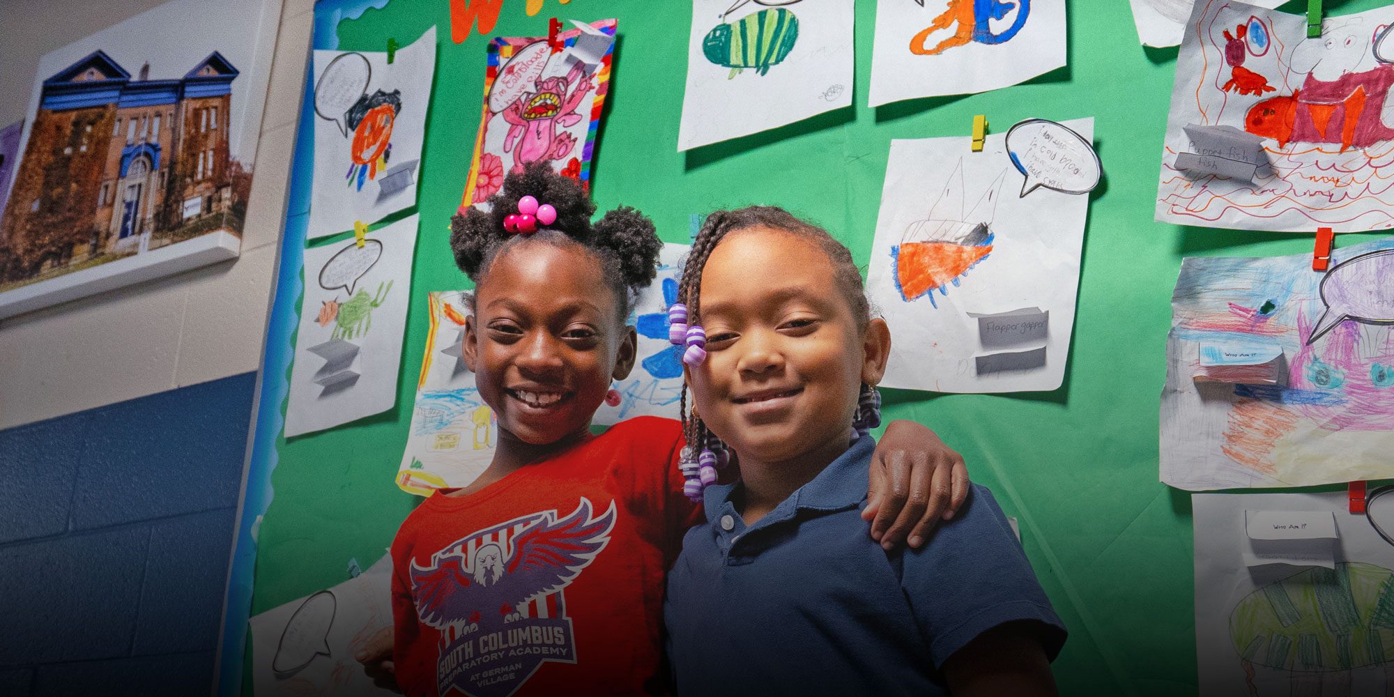 Two smiling students in front of a classroom art display.
