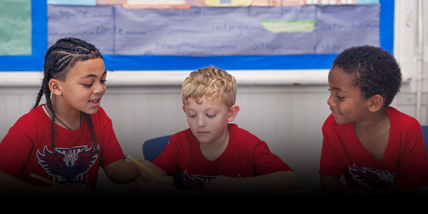 Three students in red school shirts working together at a classroom table.