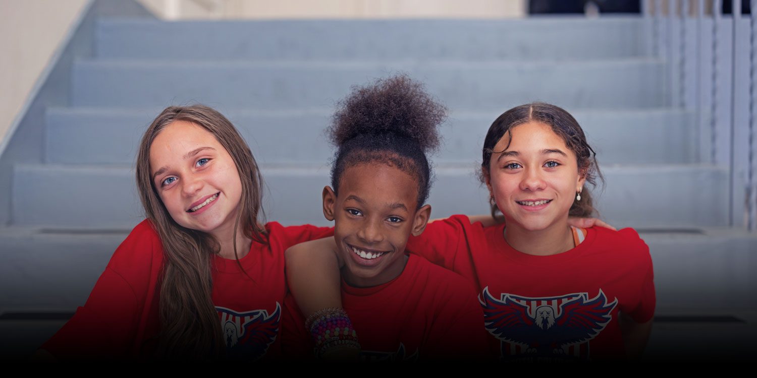 Three smiling students in red school shirts sitting together on a stairway.
